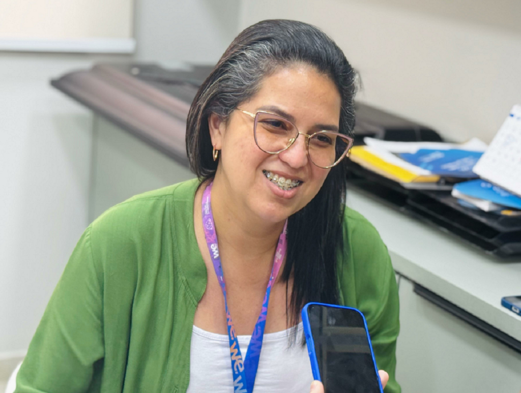 Uma mulher sorrindo. Ela usa um casaco verde e uma blusa branca. Além de estar com os seus óculos e o crachá da empresa Sebrae. Ao fundo, há uma bancada com cadernos e papéis