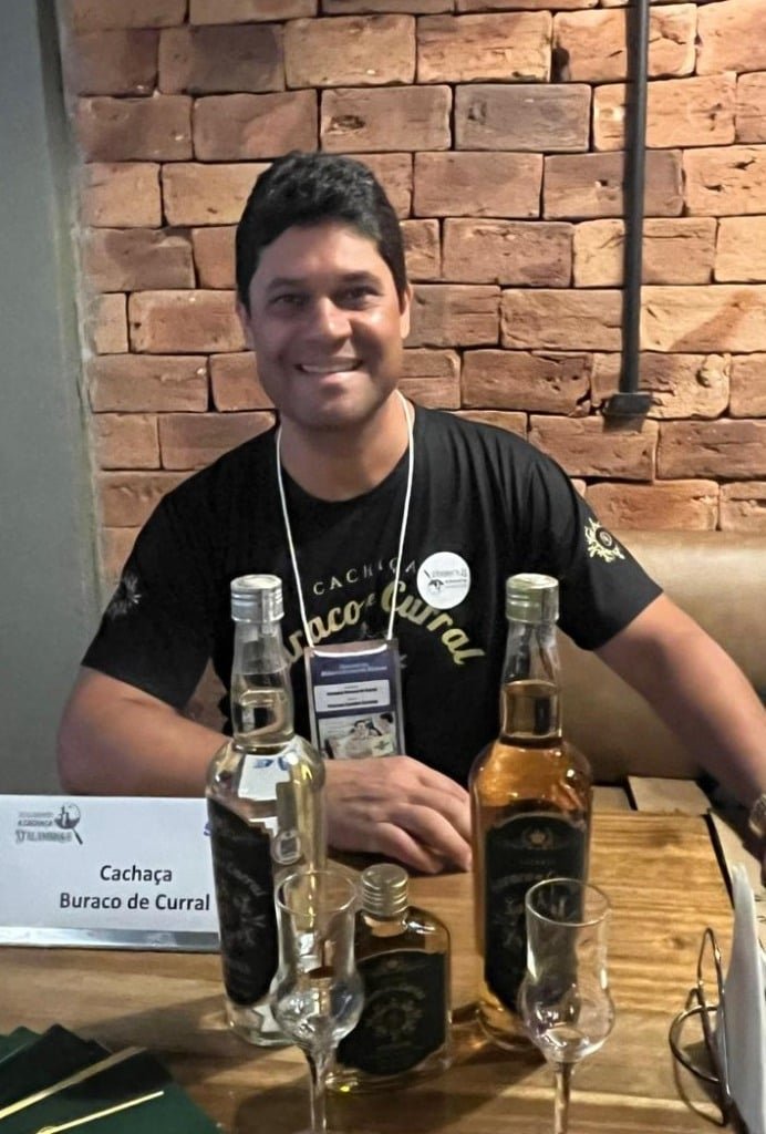 Homem com camiseta preta, sentado e à sua frente, garrafas de cachaça que ele produz