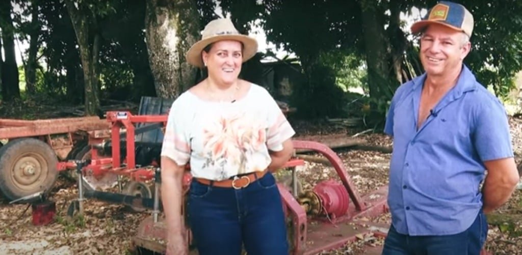 Na foto, aparece uma mulher de chapéu ao lado direito e à esquerda está um homem. Ao fundo um trator